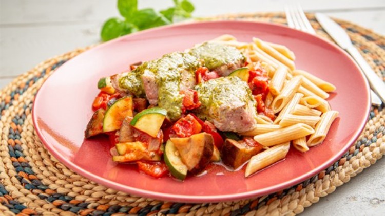 Ein roter Teller mit Penne, gebratenem Gemüse, Schweinefilet und grünem Pesto auf einem geflochtenen Untersetzer.