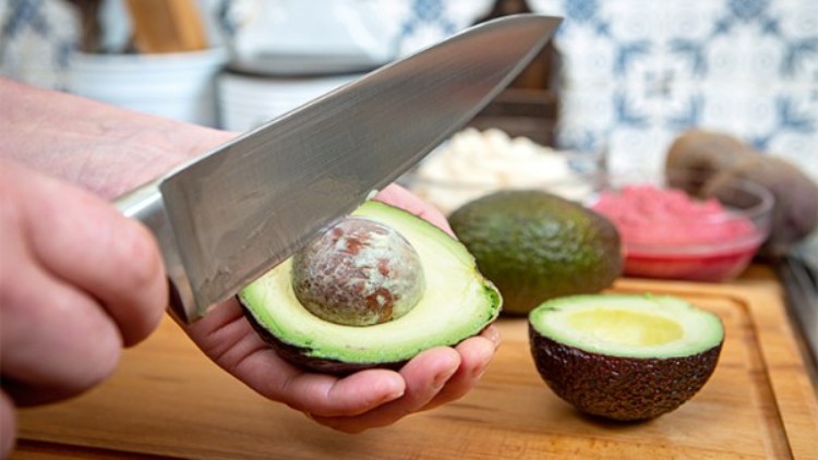 Eine halbierte Avocado wird mit einem Messer in der linken Hand gehalten und der Kern mit der Messerklinge herausgelöst.