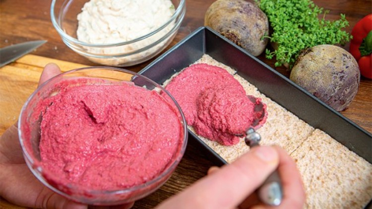 Rote Creme wird mit einem Löffel auf Brotscheiben in einer rechteckigen Backform verteilt, daneben Schalen und Gemüse.