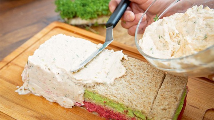 Eine rechteckige Schichttorte aus Brot mit farbigen Aufstrichen wird mit einer cremigen Masse bestrichen, Kräuter im Hintergrund.