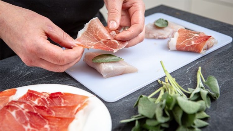 Hände wickeln eine Scheibe Schinken um Fischfilet mit Salbeiblatt auf einem Schneidebrett, daneben frische Salbeiblätter.