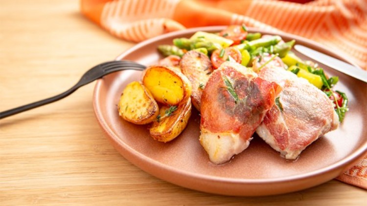 Gebratene Kartoffeln, mit Schinken umhüllter Fisch und Gemüse wie Spargel und Tomaten auf einem rosa Teller mit Besteck.