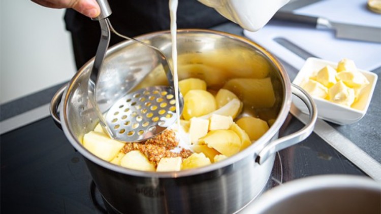 Kartoffeln, Butter und Senf in einem Edelstahltopf, darüber wird Milch aus einem weißen Gefäß gegossen, Kartoffelstampfer sichtbar.
