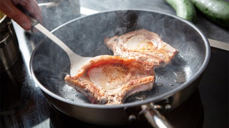 Zwei gebratene Schweinekoteletts in einer dampfenden Pfanne, eine Hand hält einen Pfannenwender, im Hintergrund Gurken.