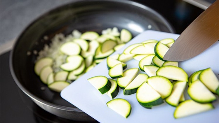 Eine Pfanne mit Zwiebeln auf einem Herd, daneben wird geschnittene Zucchini von einem Schneidebrett hineingeschoben.