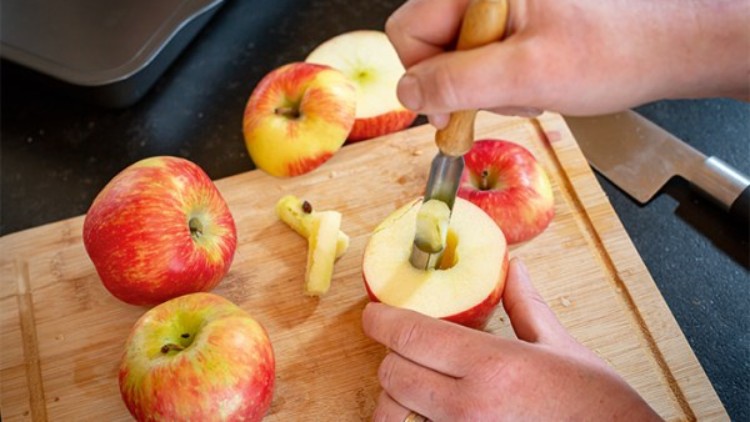 Eine Hand entkernt einen halbierten roten Apfel mit einem Apfelentkerner auf einem Holzbrett.