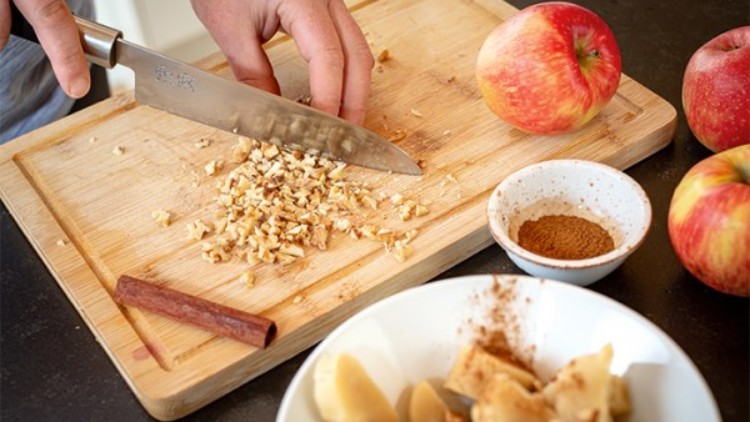 Eine Hand hackt Nüsse mit einem Messer auf einem Holzbrett, daneben Äpfel, Zimtstange, Gewürzschale und Obststückchen.
