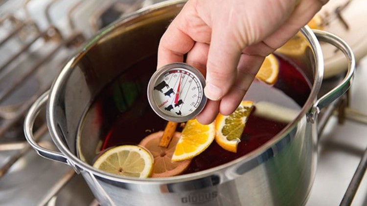 Eine Hand hält ein Thermometer über einen Topf mit heißem Getränk, Zitrusscheiben und Zimtstange auf einem Herd.