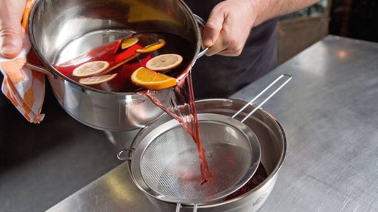 Eine Person gießt roten Fruchtpunsch mit Orangen- und Zitronenscheiben durch ein Sieb in eine Metallschüssel.