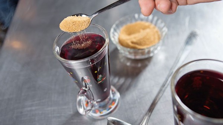 Ein Teelöffel brauner Zucker wird über ein Glas mit heißem, dunklem Getränk geschüttet, im Hintergrund steht eine Zuckerschale.
