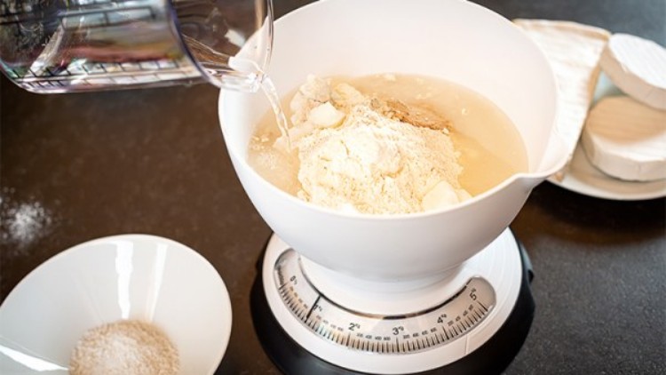 Eine weiße Schüssel mit Mehl auf einer Küchenwaage, daneben Wasser, ein Teller mit Salz und Käse auf schwarzer Arbeitsfläche.