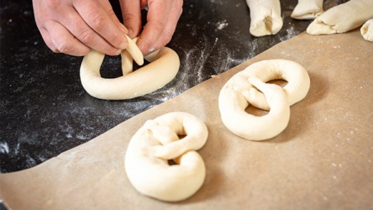 Hände formen aus Teig eine Brezel, daneben liegen weitere Brezeln auf einer bemehlten Arbeitsfläche und Backpapier.