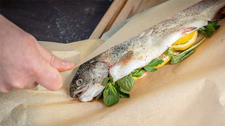 Eine Hand wickelt einen mit Zitronenscheiben, Minze und Kräutern gefüllten Fisch in braunes Backpapier.