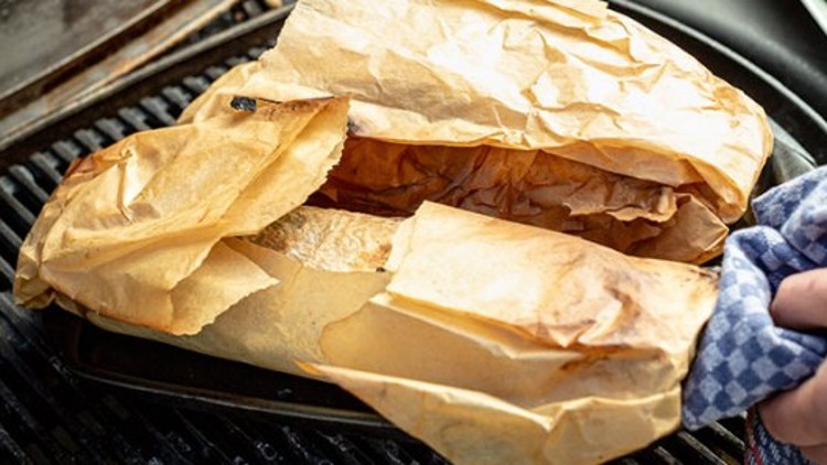 Ein Backblech mit braunem Backpapier, das sich teilweise öffnet, wird mit einem blauen Geschirrtuch gehalten.