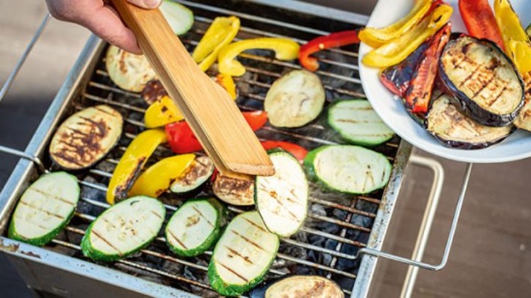 Ein kleiner Grill mit Zucchini, Paprika und Auberginenscheiben, eine Hand legt Gemüse vom Teller mit einem Holzspatel darauf.