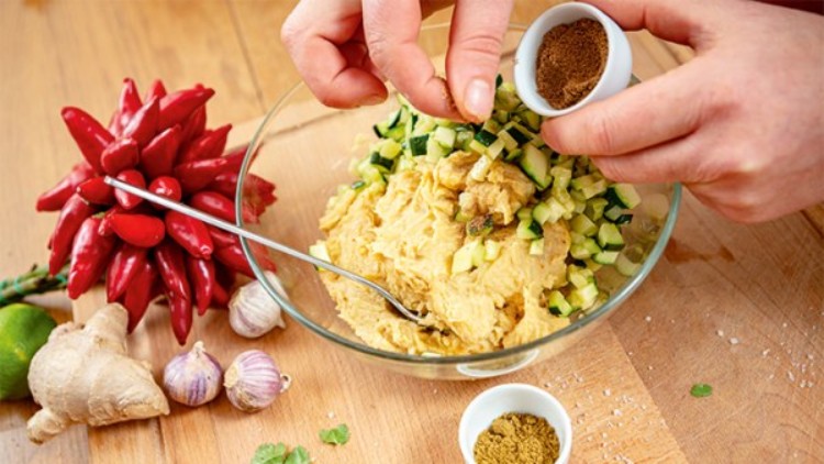 Hände geben Gewürz aus einem kleinen weißen Schälchen in eine Schüssel mit Hummus, Zucchiniwürfeln und einem Löffel.