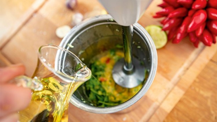 Handmixer püriert grüne und rote Zutaten in einem metallischen Gefäß, während aus einem Glas Öl hinzugegossen wird.