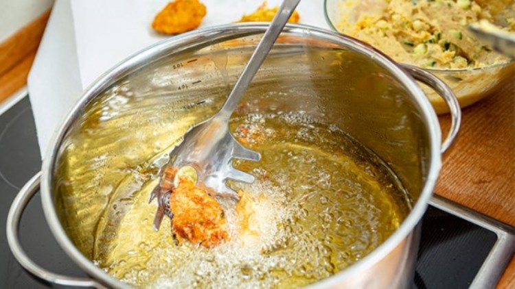 Ein Metalltopf mit heißem Öl enthält eine Zange, die ein frittiertes Stück Teig heraushebt, daneben steht eine Schüssel mit Teig.