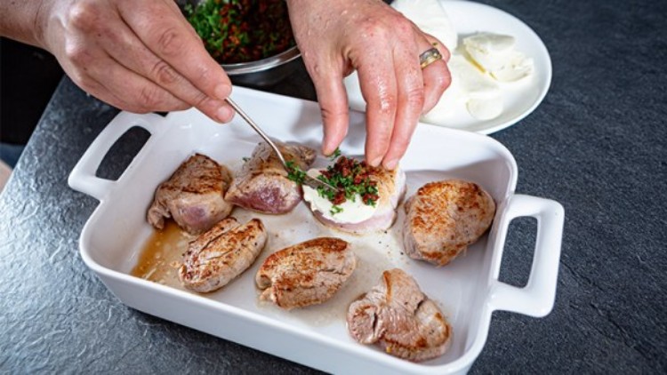 Person legt mit einem Löffel Kräuter-Mischung auf ein Stück Fleisch in einer weißen Auflaufform, daneben liegt Mozzarella.