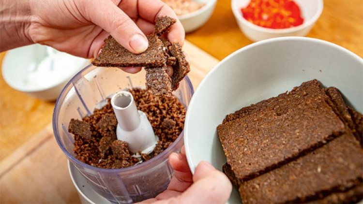 Zwei Hände zerbröseln dunkles Brot in einen Zerkleinerer, daneben eine Schüssel mit Brotscheiben und weitere Schüsseln im Hintergrund.
