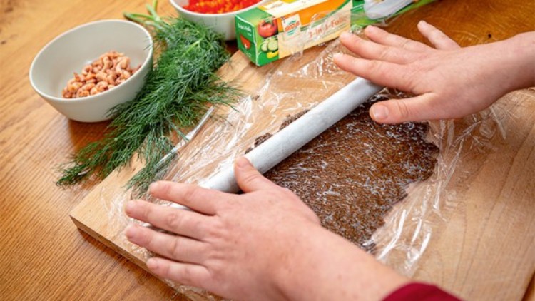 Hände rollen zwischen Frischhaltefolie eine dunkle Masse auf einem Holzbrett aus, daneben Dill, Garnelen und Frischhaltefolie.