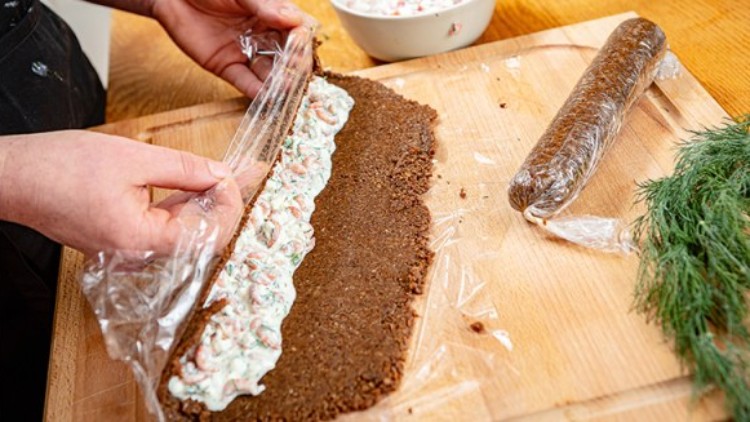 Eine Hand rollt mit Frischhaltefolie eine dunkle Teigmasse, gefüllt mit heller Creme und Kräutern, auf einem Holzbrett.