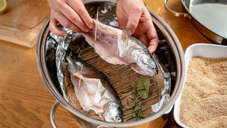 Zwei ganze Fische liegen in einem Metalltopf mit Kräutern auf einem Rost, eine Person hält einen geöffneten Fisch über dem Topf.
