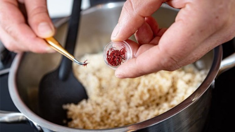 Eine Hand hält Safranfäden in einer Pinzette über einen Topf mit gekochtem Quinoa, daneben ein offenes Gefäß mit Safran.
