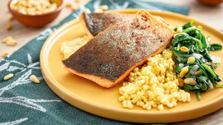 Gebratener Lachs mit knuspriger Haut auf Hirse, Spinat und gerösteten Pinienkernen auf einem gelben Teller.