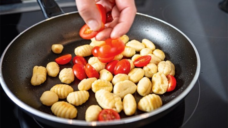 Hand fügt halbierte Kirschtomaten zu goldbraun gebratenen Gnocchi in einer schwarzen Pfanne auf einer Herdplatte hinzu.