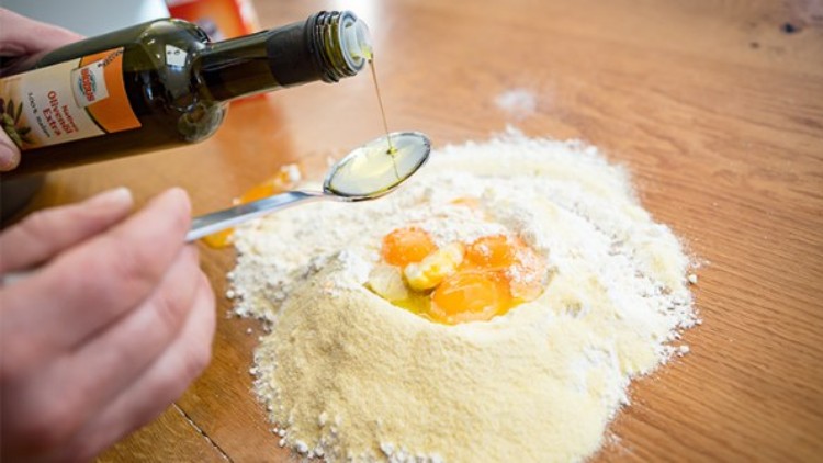 Mehlhügel mit Eiern und Butter auf Holzuntergrund, dazu wird Olivenöl aus einer Flasche auf einen Löffel gegossen.