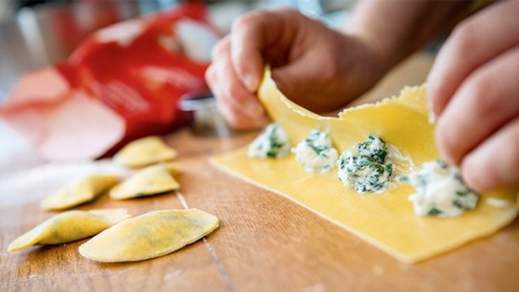 Eine Person füllt Teigtaschen mit grün-weißer Spinat-Ricotta-Mischung auf einem Holzuntergrund.