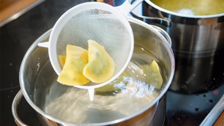 Ein weißes Küchensieb hebt gelbe Teigtaschen aus einem Topf mit kochendem Wasser, weitere Töpfe stehen auf dem Herd.