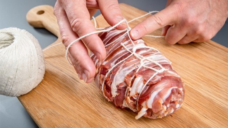 Hände binden mit weißer Schnur einen mit Speck umwickelten Fleischrollbraten auf einem Holzbrett, daneben liegt ein Garnknäuel.