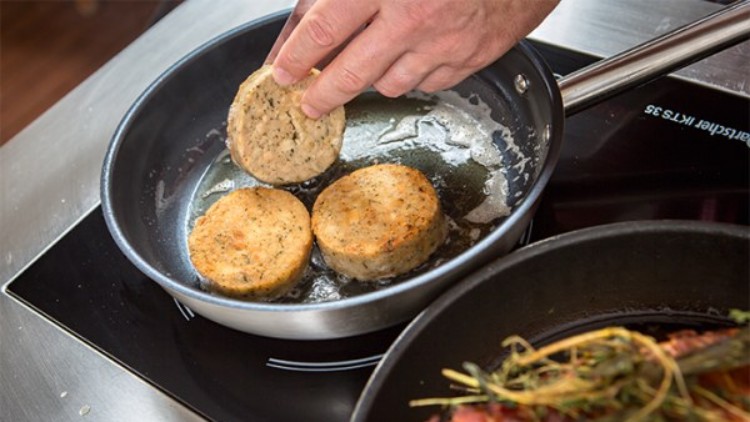Hand legt vegetarischen Bratling in eine Pfanne mit zwei weiteren Bratlingen auf einem Herd, daneben liegt eine Pfanne mit Garnelen.