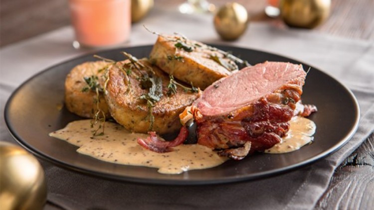 Ein Teller mit rosa gebratenem Fleisch, zwei Serviettenknödeln und heller Senfsauce, garniert mit frischen Kräutern.