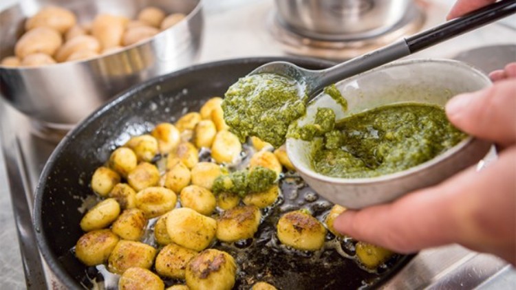 Gebratene Gnocchi in einer Pfanne, dazu wird mit einem Löffel grüne Pesto aus einer kleinen Schale hinzugegeben.