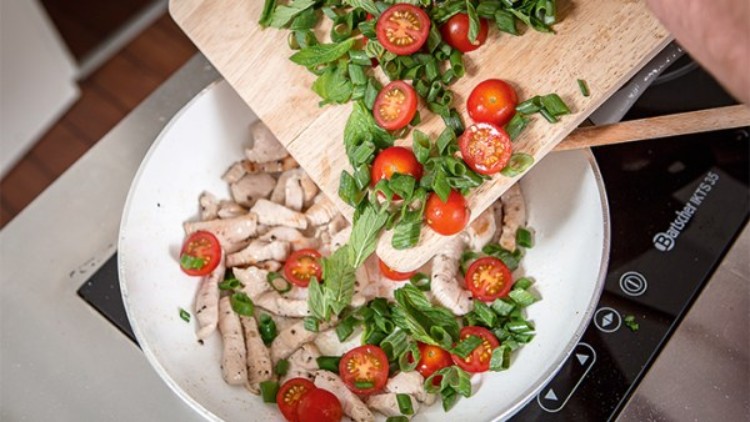 Geschnittene Kirschtomaten und Frühlingszwiebeln werden von einem Holzbrett in eine Pfanne mit gebratenem Fleisch gegeben.