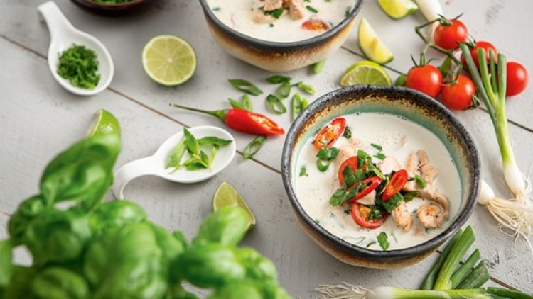 Zwei Schalen Suppe mit Kokosmilch, Gemüse und Chili, dekoriert mit frischen Kräutern, umgeben von Tomaten, Limetten und Frühlingszwiebeln.