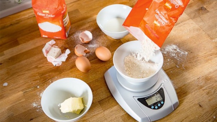Auf einer Küchenwaage werden 250 Gramm Mehl abgewogen, daneben liegen Zucker, Butter, Eier und eine Schüssel bereit.