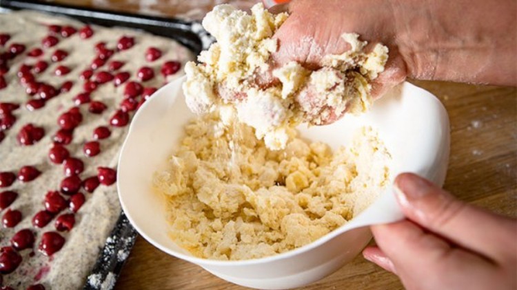 Eine Hand formt Streuselteig in einer weißen Schüssel, im Hintergrund ein Blech mit Kirschen auf Kuchen.