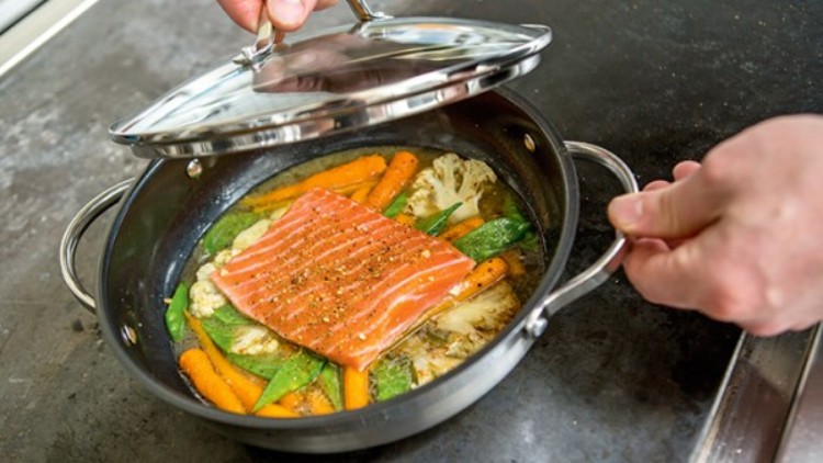 Lachsfilet mit Gewürzen liegt auf Gemüse wie Möhren, Blumenkohl und Zuckerschoten in einer Pfanne, die gerade geschlossen wird.