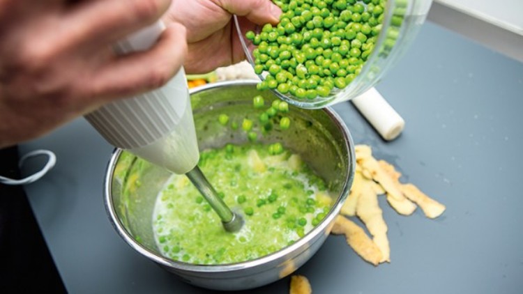 Eine Person püriert grüne Erbsen in einer Metallschüssel mit einem Handmixer, daneben Schalenreste auf einer grauen Fläche.