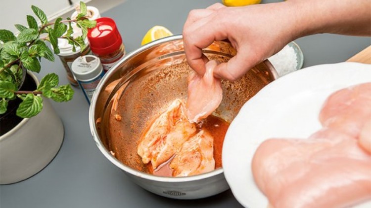 Eine Hand legt mariniertes Hähnchenfilet aus einer Schüssel mit Marinade auf einen Teller, daneben frische Minze und Gewürze.