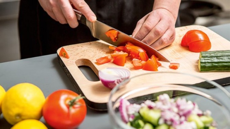 Hände schneiden Tomaten auf einem Holzbrett, daneben liegen Zitrone, Gurke, Tomaten und eine Glas-Schüssel mit Salatzutaten.
