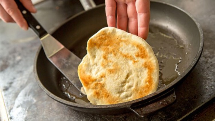 Ein frisch gebackenes, goldbraunes Fladenbrot wird in einer schwarzen Pfanne mit einem Pfannenwender gewendet.