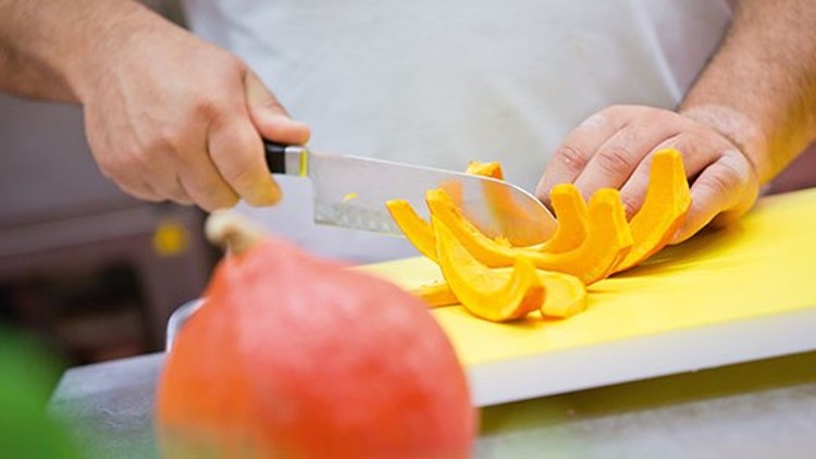 Eine Person schneidet mit einem Messer orangefarbenes Kürbisfleisch auf einem gelben Schneidebrett.