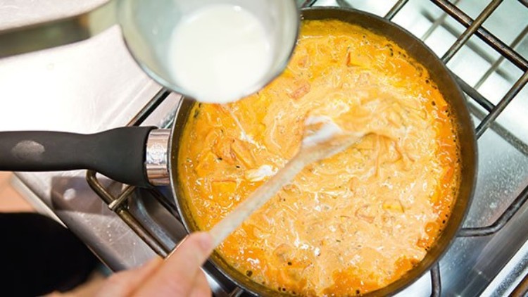 Eine orangene Soße wird in einer Pfanne auf dem Herd mit weißer Flüssigkeit und einem Holzlöffel verrührt.