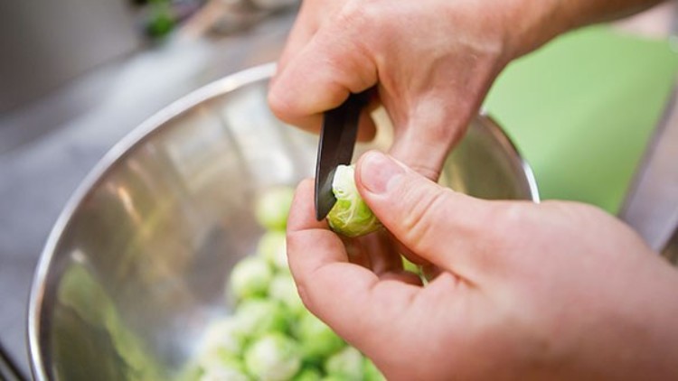Hände schneiden mit einem Messer den Strunk von frischem Rosenkohl über einer silbernen Schüssel ab.