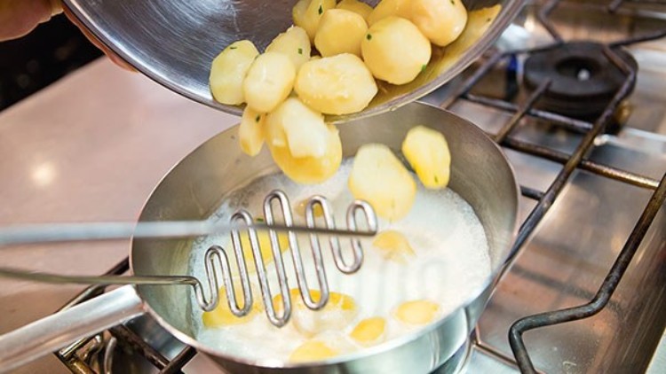 Pellkartoffeln werden aus einer Metallschüssel in einen Topf mit Milch auf einem Herd mit Gasflamme gegeben, Stampfer liegt im Topf.